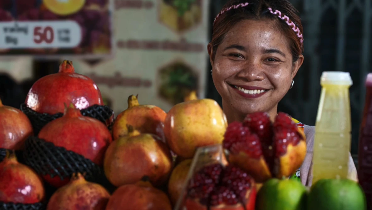 Smiling vendor at a fruit market stand displaying pomegranates and other fresh produce.