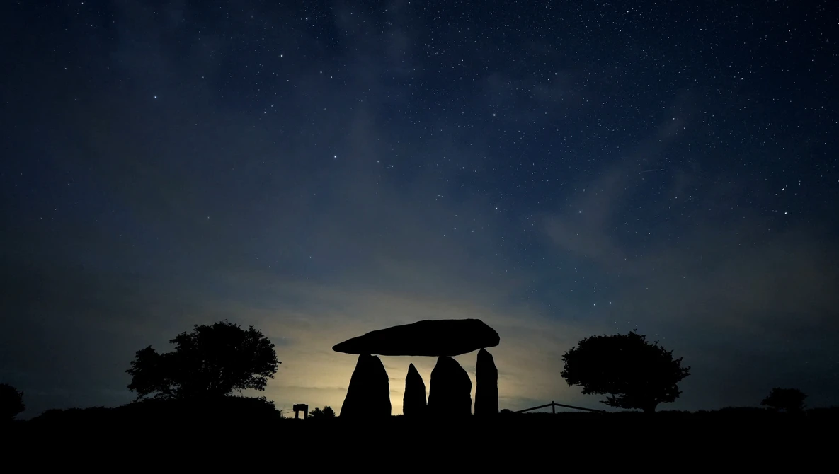 Silhouette of Stonehenge monument against a twilight sky with scattered stars and trees on the horizon.