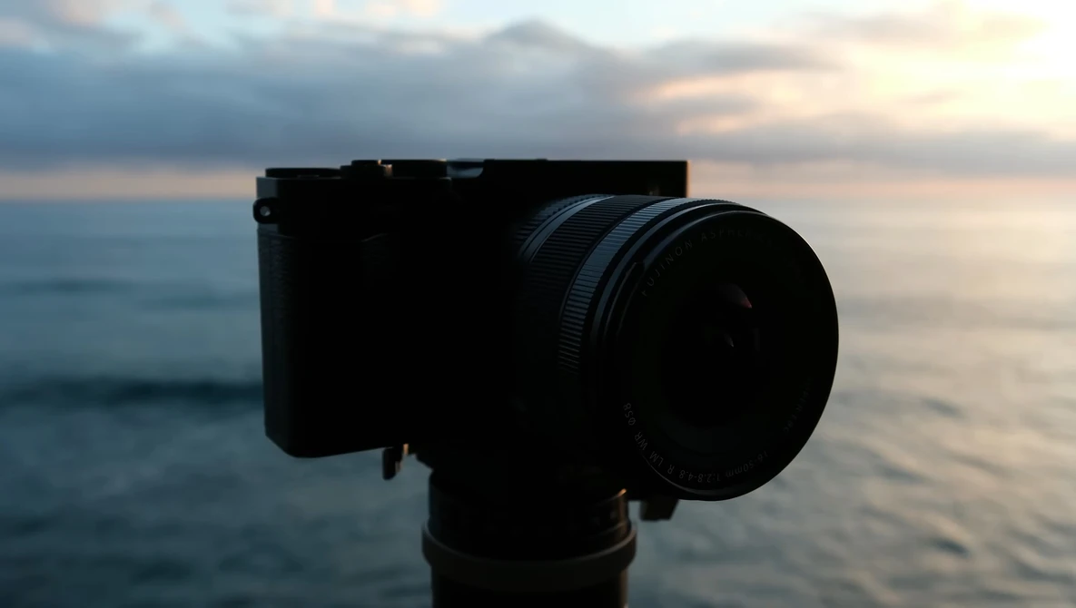 Compact camera with telephoto lens mounted on tripod, silhouetted against ocean and sunset sky.
