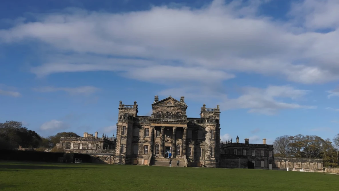 Stone castle with crenellated towers set on manicured grounds under partly cloudy sky.