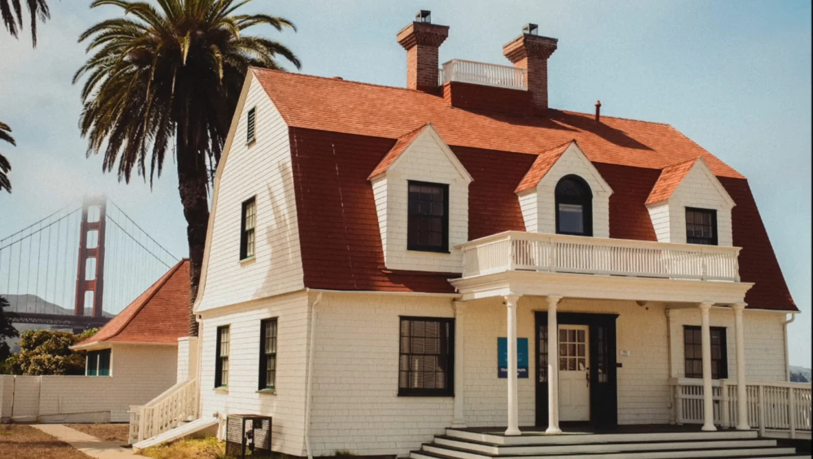 White Victorian house with red mansard roof and palm tree beside Golden Gate Bridge in background.