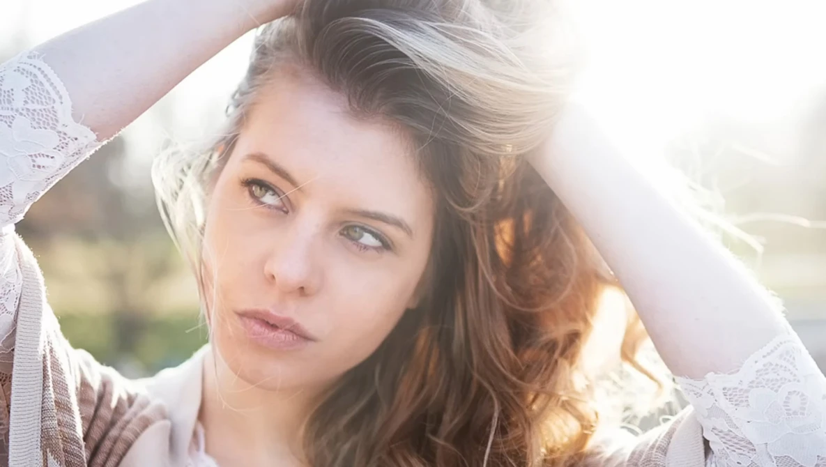 Woman with long brown hair backlit by golden sunlight, holding a white lace fabric above her head.