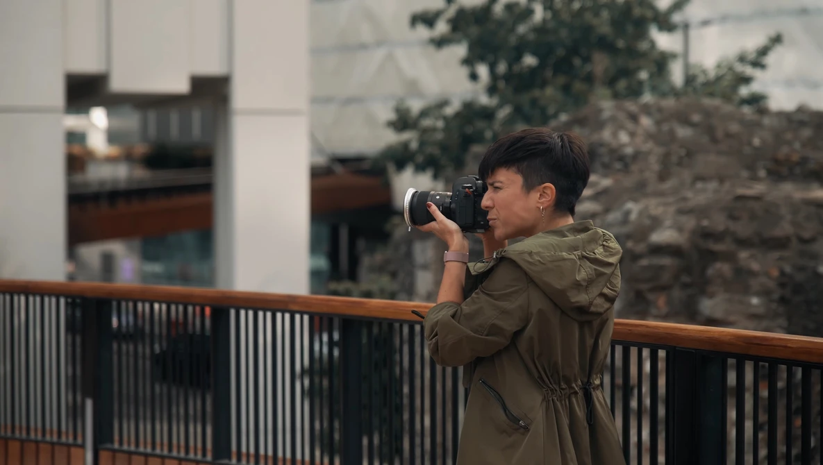 Photographer in olive jacket holding camera to eye while standing at railing overlooking urban architecture.