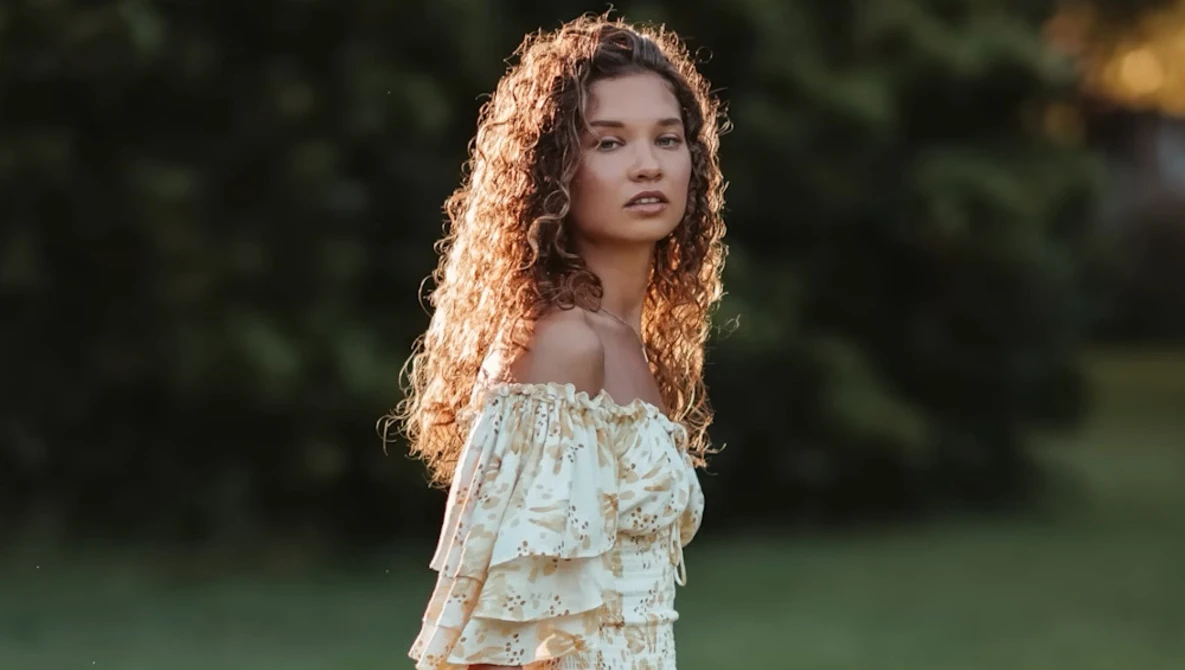 Young woman with long curly copper-toned hair wearing an off-shoulder ruffle blouse in a garden setting.