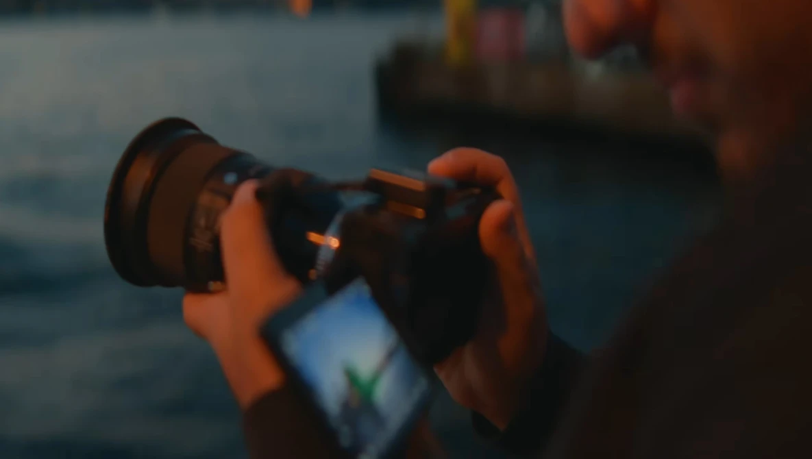 Photographer holding a camera with smartphone mounted below, capturing a sunset over water with boats in the background.