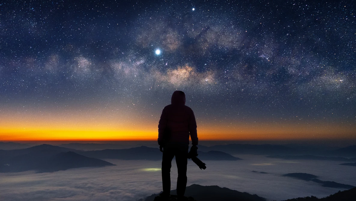 Silhouetted figure standing on a mountain peak overlooking a sea of clouds beneath a star-filled sky and Milky Way.
