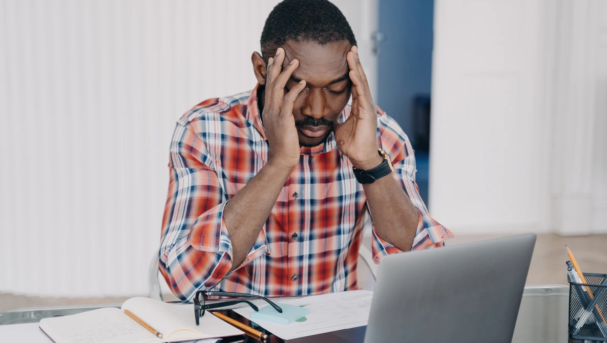Man in plaid shirt experiencing stress while working at laptop at desk.