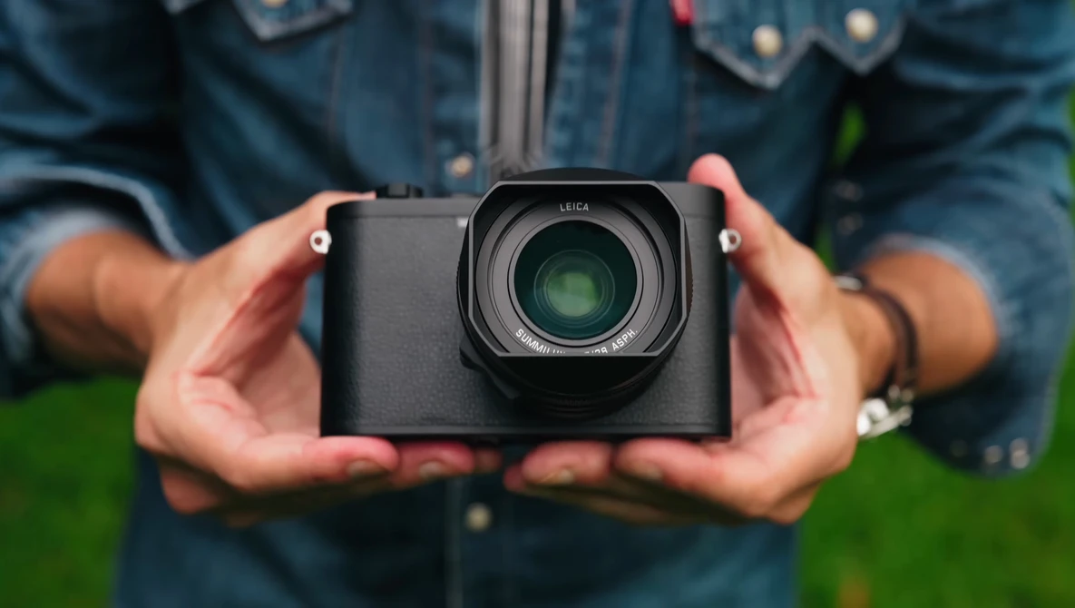 Photographer holding a compact mirrorless camera with a prime lens against a blurred green background.