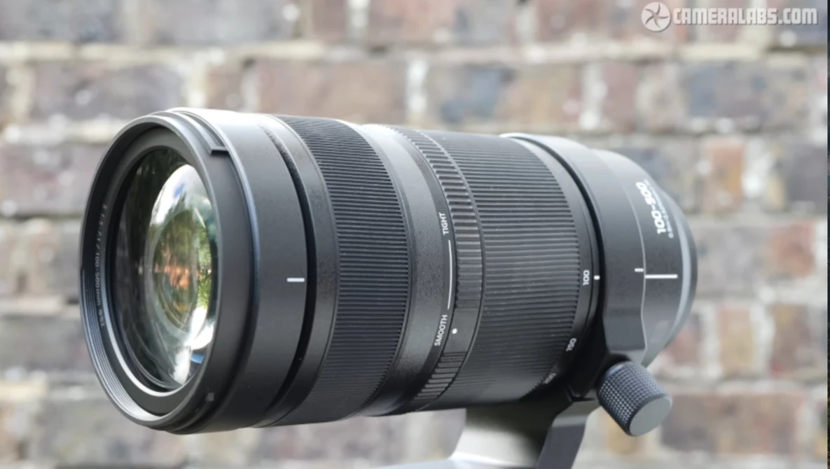 Black telephoto zoom lens mounted on support with brick wall backdrop.