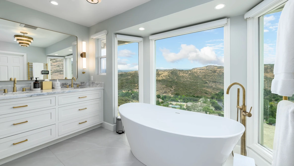 Modern bathroom with freestanding white oval tub positioned in front of large windows overlooking green hillside landscape.
