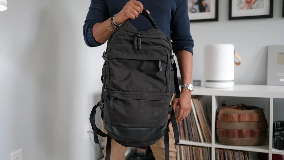 Person holding a black multi-pocket backpack with shoulder straps in an interior setting.
