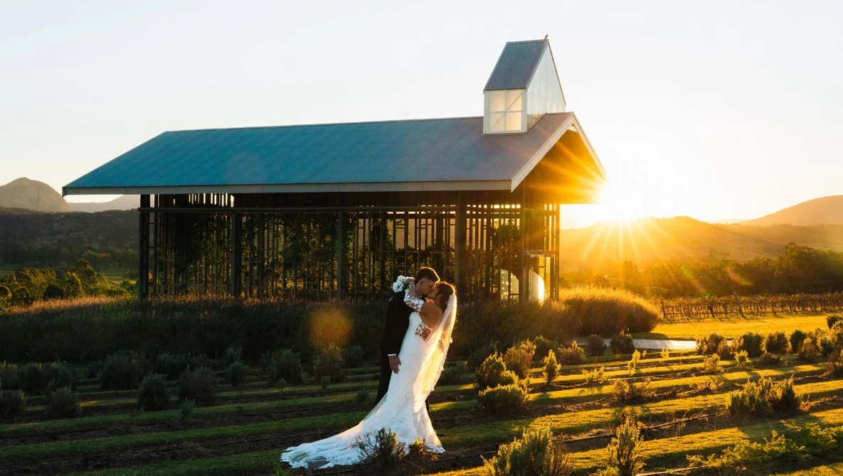 Bride in flowing white gown posing with groom at sunset in front of a modern barn structure with vineyards in the background.