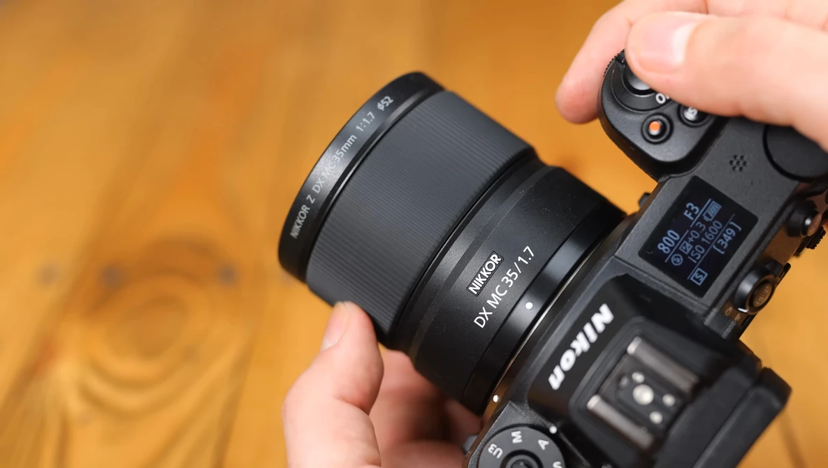 Hand holding a mirrorless camera with a prime lens attached against a warm orange background.