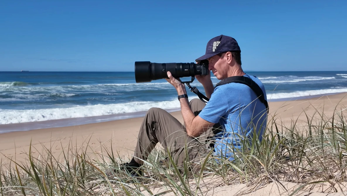 Photographer seated in beach grass using a telephoto lens to capture the ocean and shoreline.
