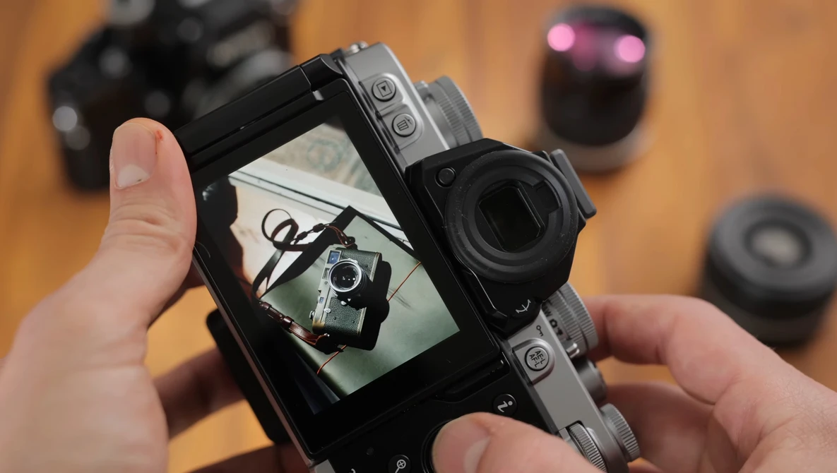 Hands holding a mirrorless camera displaying its rear LCD screen with a self-portrait of the photographer holding the same camera.