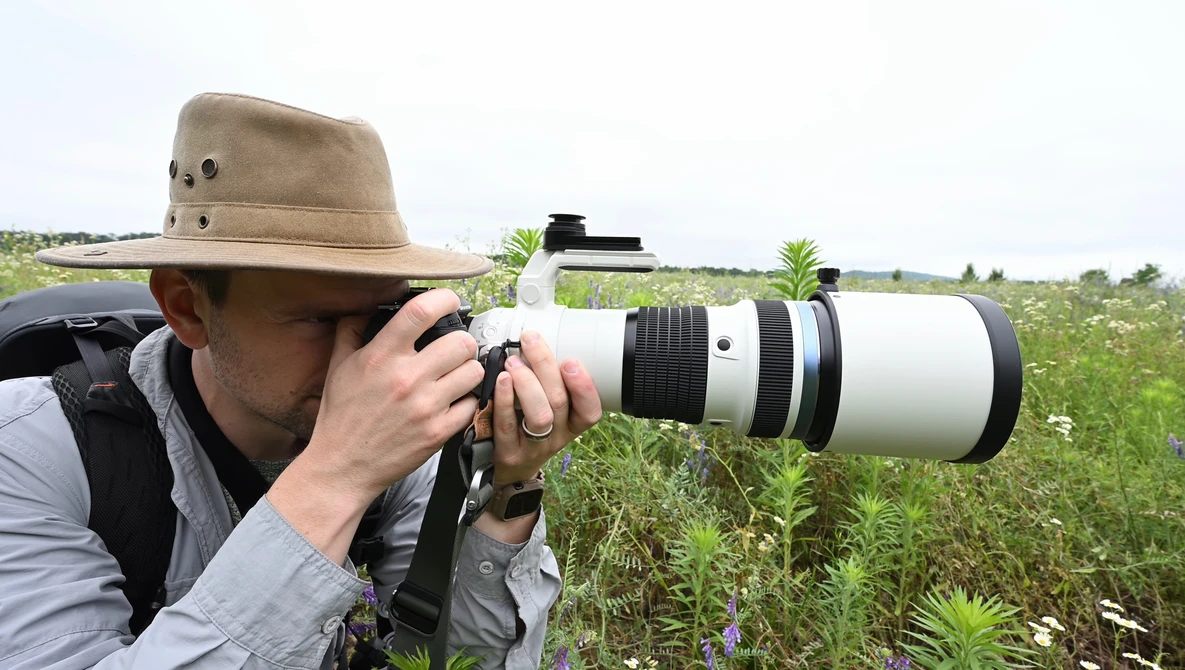 Photographer in tan hat using a white telephoto lens in an overgrown field.