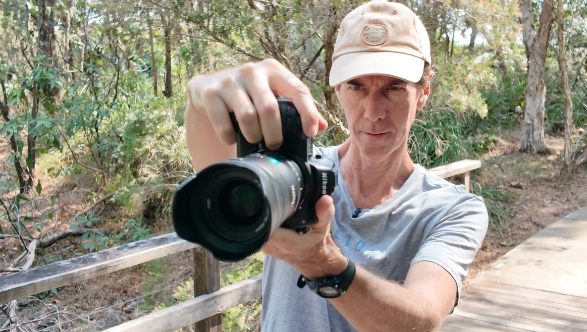 Photographer holding a DSLR with telephoto lens pointed toward camera in a wooded forest setting.
