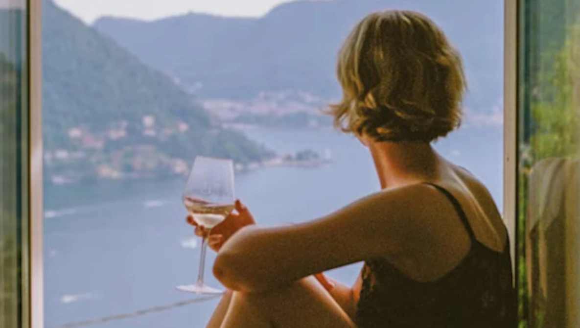 Woman holding a wine glass overlooking a mountainous lake landscape from an interior vantage point.