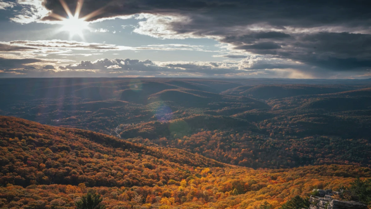 Autumn forest landscape with layered mountains beneath dramatic sky with sun rays.