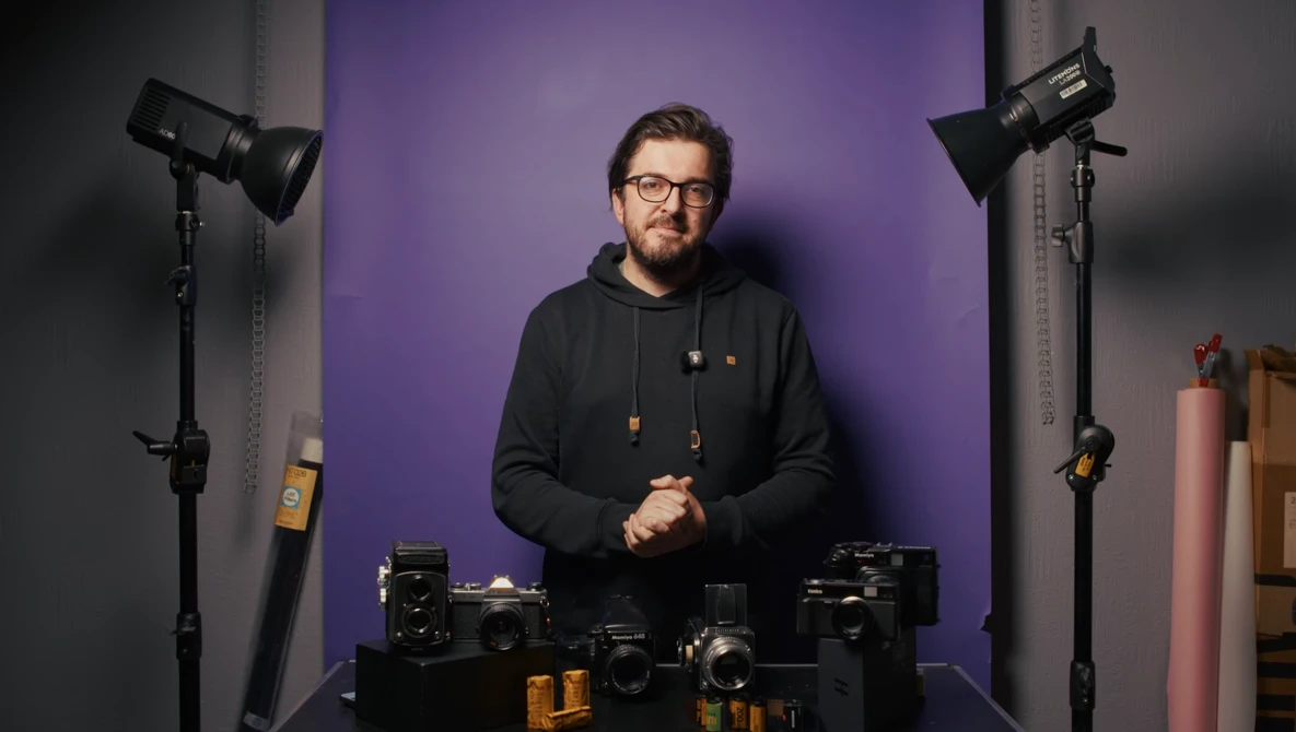 Photographer in black hoodie standing at desk with camera equipment and professional studio lighting setup.
