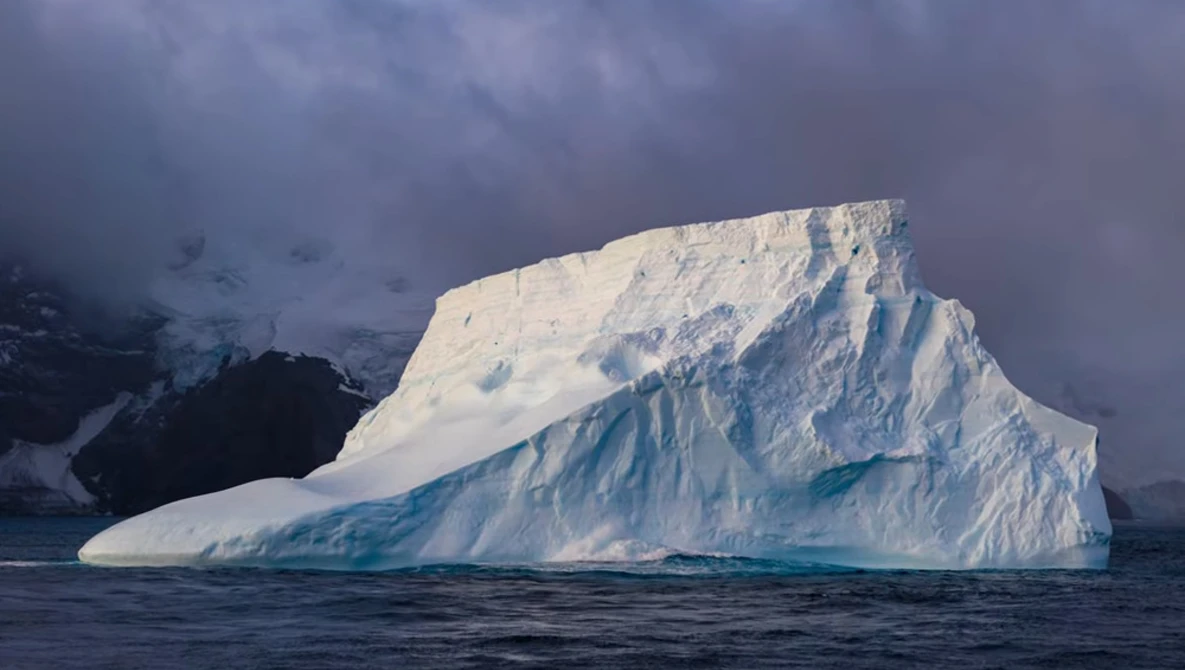 Large tabular iceberg with steep blue-tinted faces floating in dark Antarctic waters.