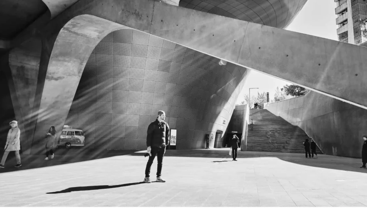 Man standing in sunlit concrete plaza beneath curved architectural structure with dramatic shadows.