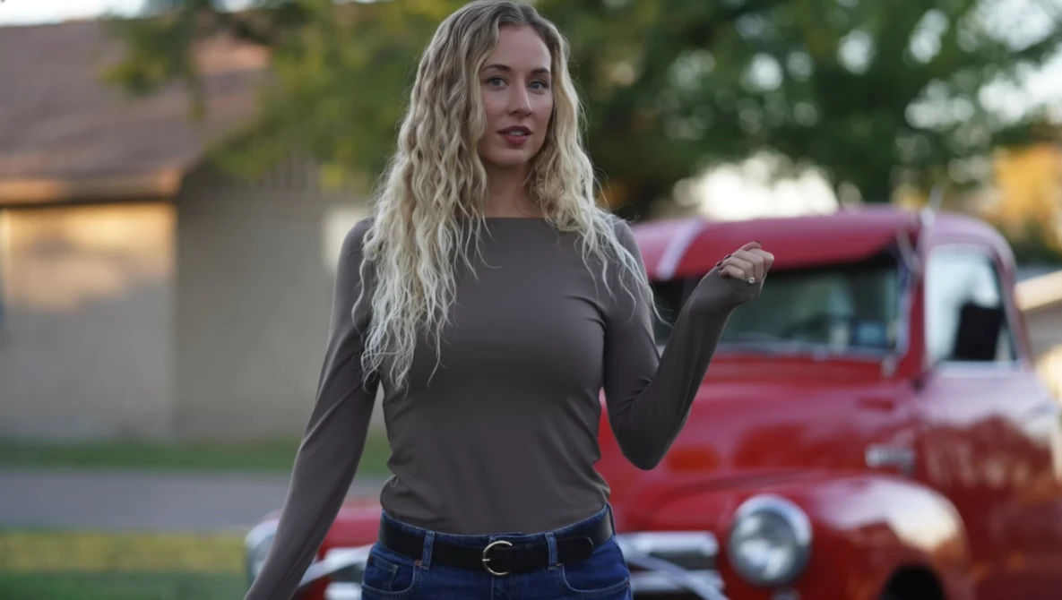 Woman with long blonde wavy hair wearing a dark long-sleeve shirt and jeans, standing outdoors with a vintage red truck blurred in the background.