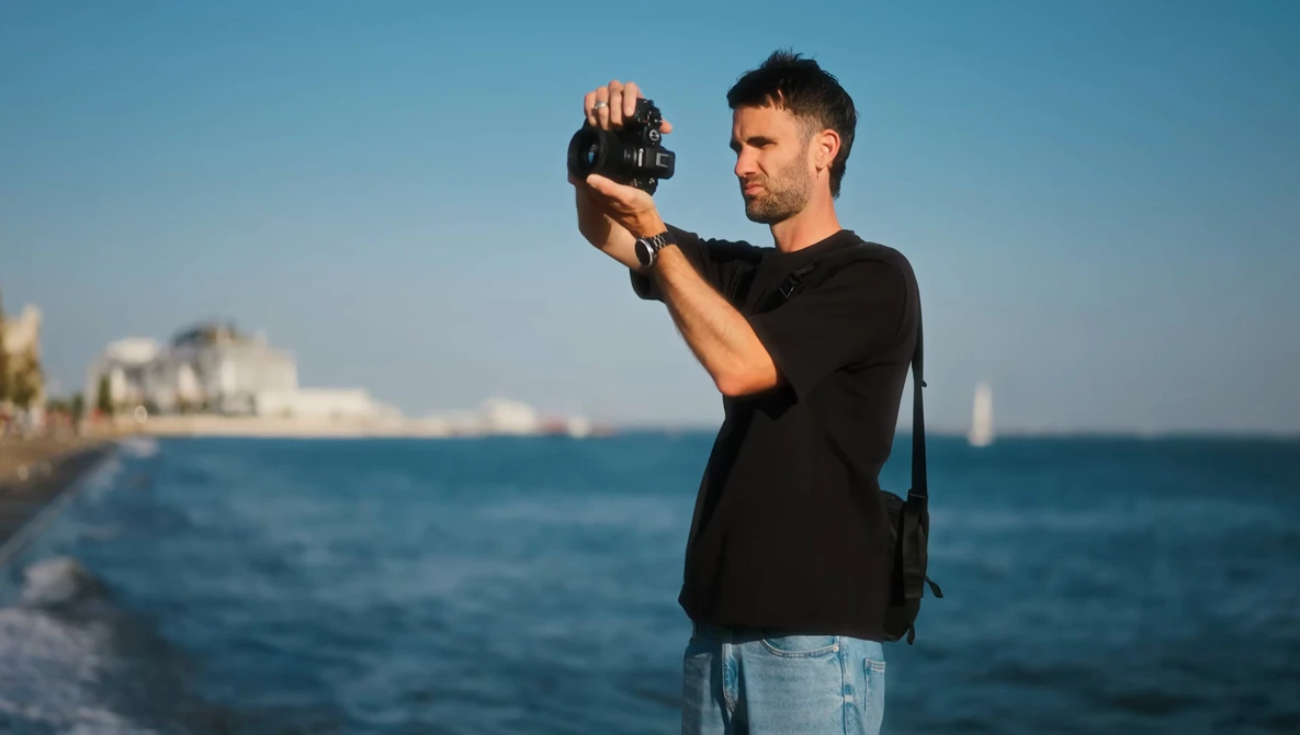 Man in black shirt holding a compact camera up to his face while standing on a rocky beach with clear blue ocean and coastal buildings in the background.