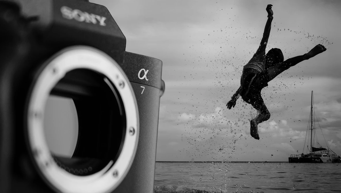 Sony α7 camera body in foreground with silhouetted person diving into water near sailboat in background, black and white.