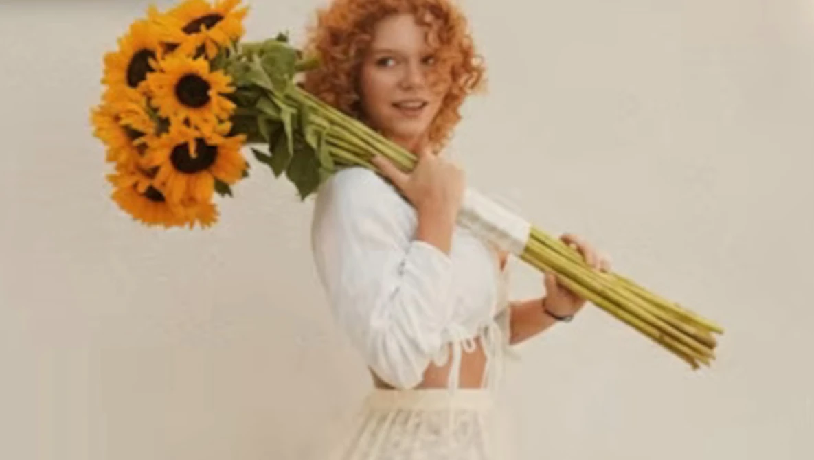 Woman with curled reddish-blonde hair holding a large bundle of yellow sunflowers over her shoulder against a neutral background.