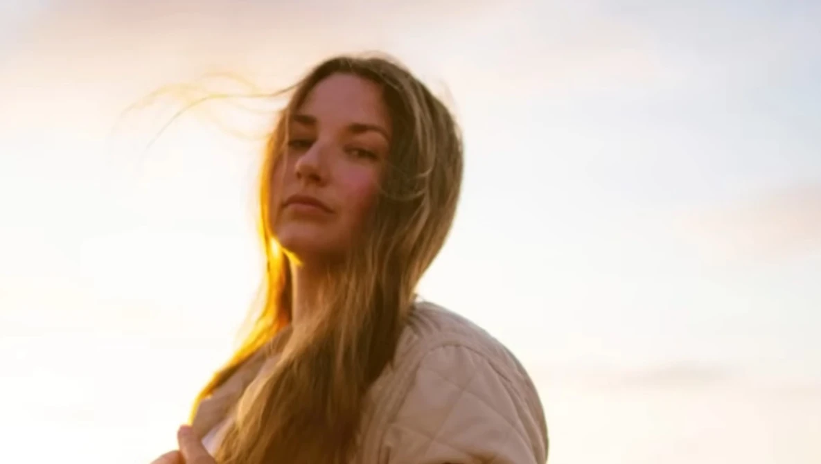 Woman with long blonde hair backlit by warm golden sunset light against a soft, overcast sky.