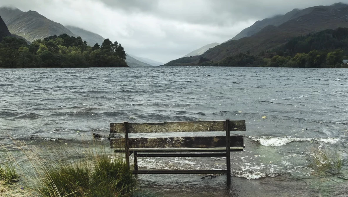 Weathered wooden bench partially submerged in calm lake water, facing mountains under overcast sky.