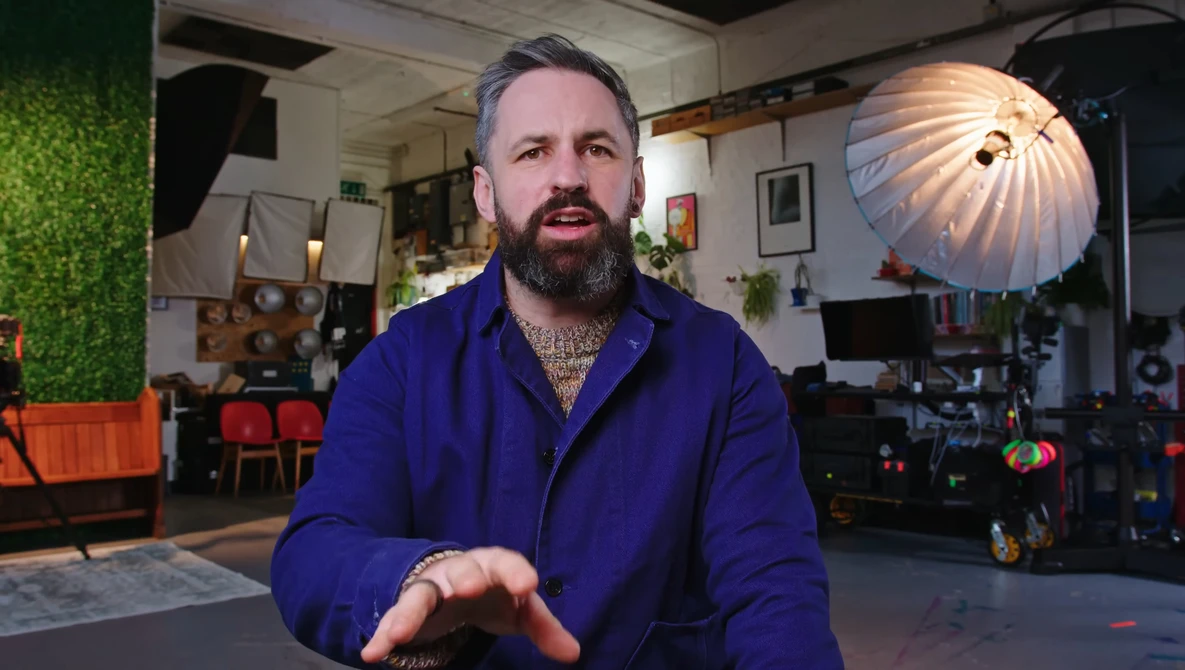Man with beard wearing blue jacket speaks directly to camera in professional studio setting.