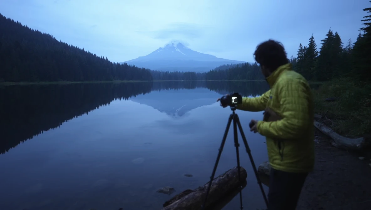 Photographer with tripod-mounted camera at dawn overlooking a calm lake with a snow-capped mountain peak reflected in the water.
