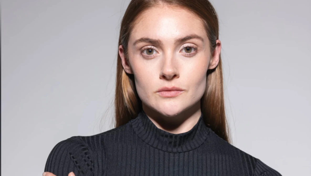 Portrait of a young woman with long brown hair wearing a dark turtleneck against a neutral gray background.