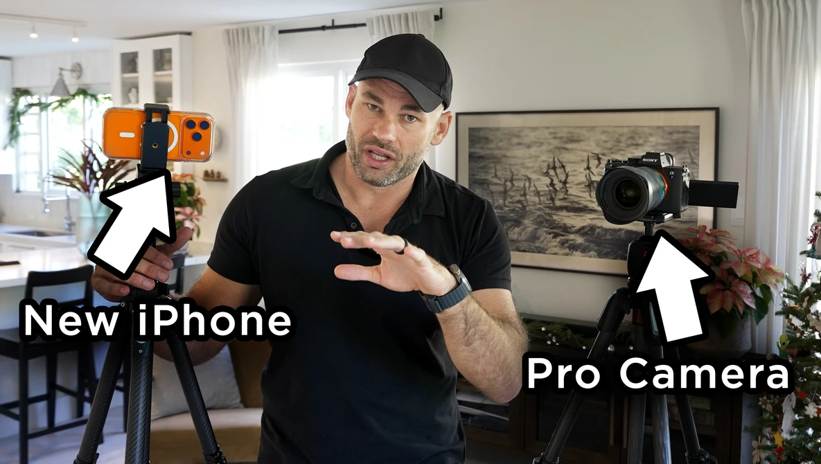 Content creator in black cap and shirt gestures between a smartphone on a tripod and a professional cinema camera, both labeled with white arrows in a bright home studio.