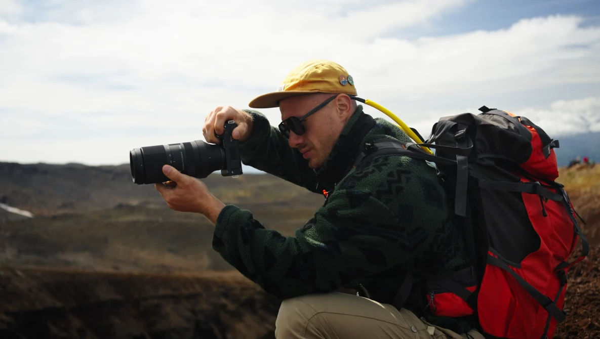 Photographer in yellow hard hat and backpack crouches while composing a shot with a telephoto lens in an arid landscape.