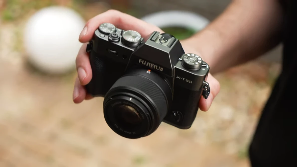 Hand holding a Fujifilm X-T30 mirrorless camera with attached lens outdoors.