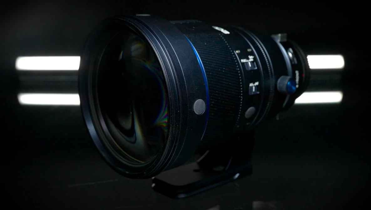 Wide-angle camera lens with blue focus ring, backlit by studio lights against dark background.