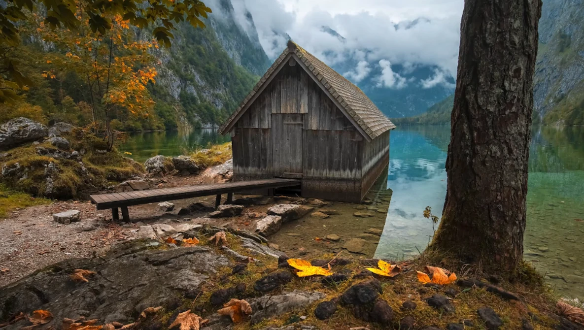 Weathered boathouse on an alpine lake shore with forested mountains and turquoise water.