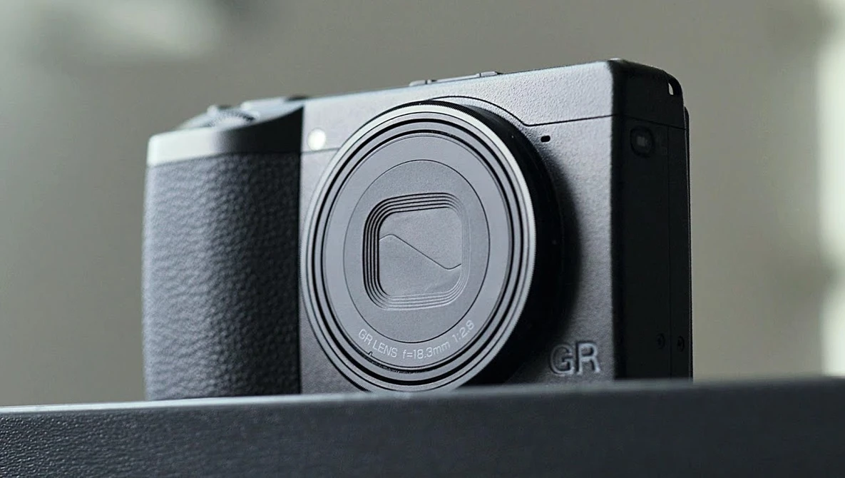Ricoh GR compact camera with silver top plate and black grip, shown in three-quarter view against neutral background.