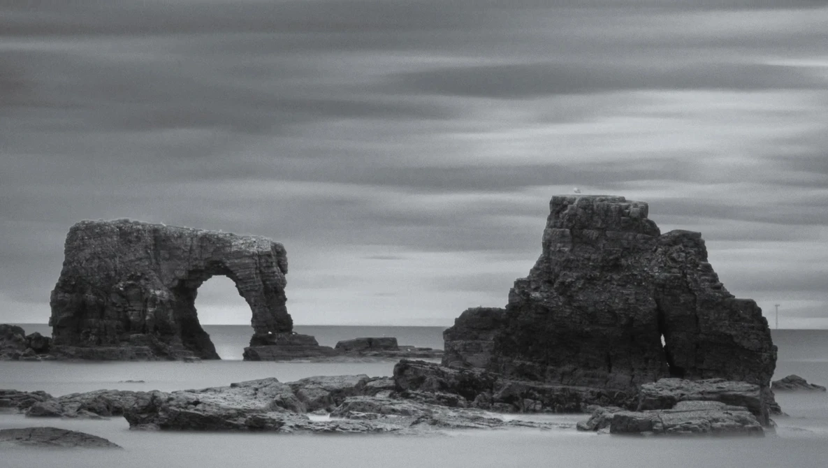 Rock formations with natural arch rising from shallow ocean water under dramatic cloudy sky.