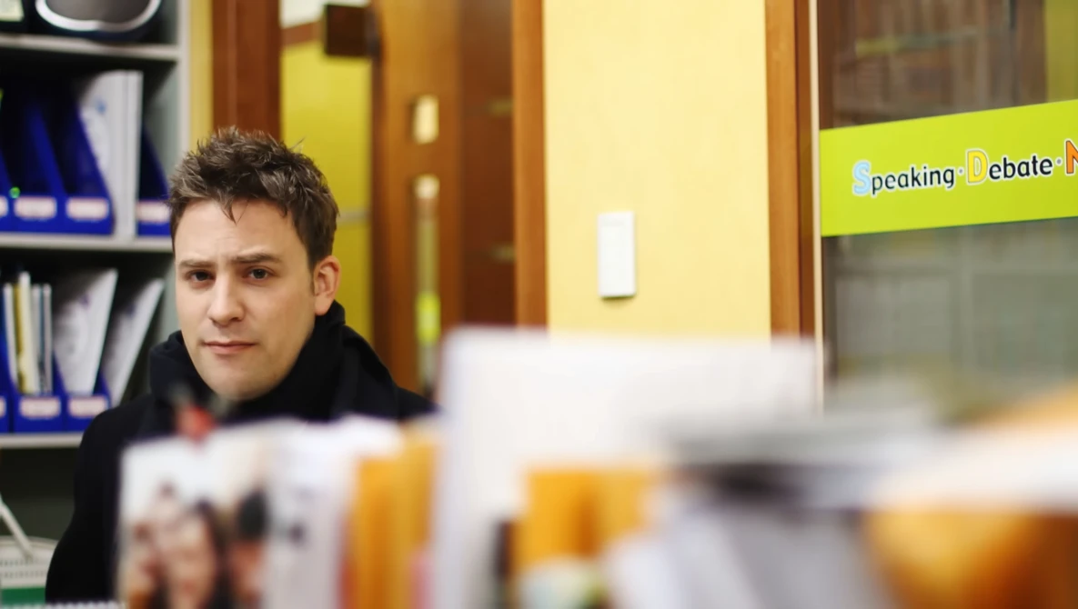 Young man in dark jacket looking at camera in bright interior space with yellow walls and shelving.