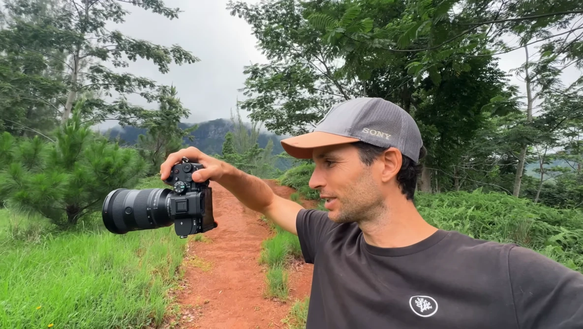 Photographer in gray cap holds mirrorless camera up to eye while standing on red dirt path surrounded by lush green vegetation and distant mountains.