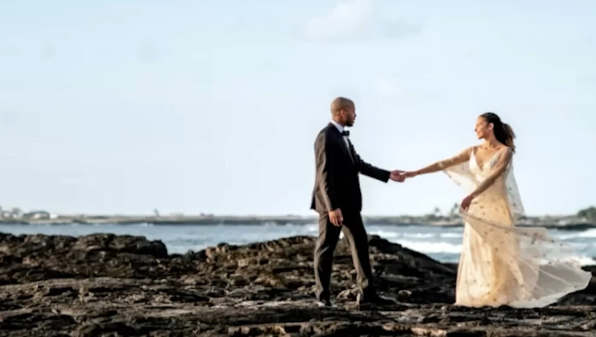 Couple holding hands on rocky beach with ocean and sky in background.