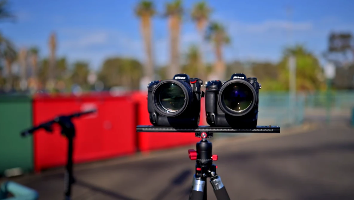 Two cinema cameras mounted on a rig against a blurred outdoor background with palm trees and red equipment.