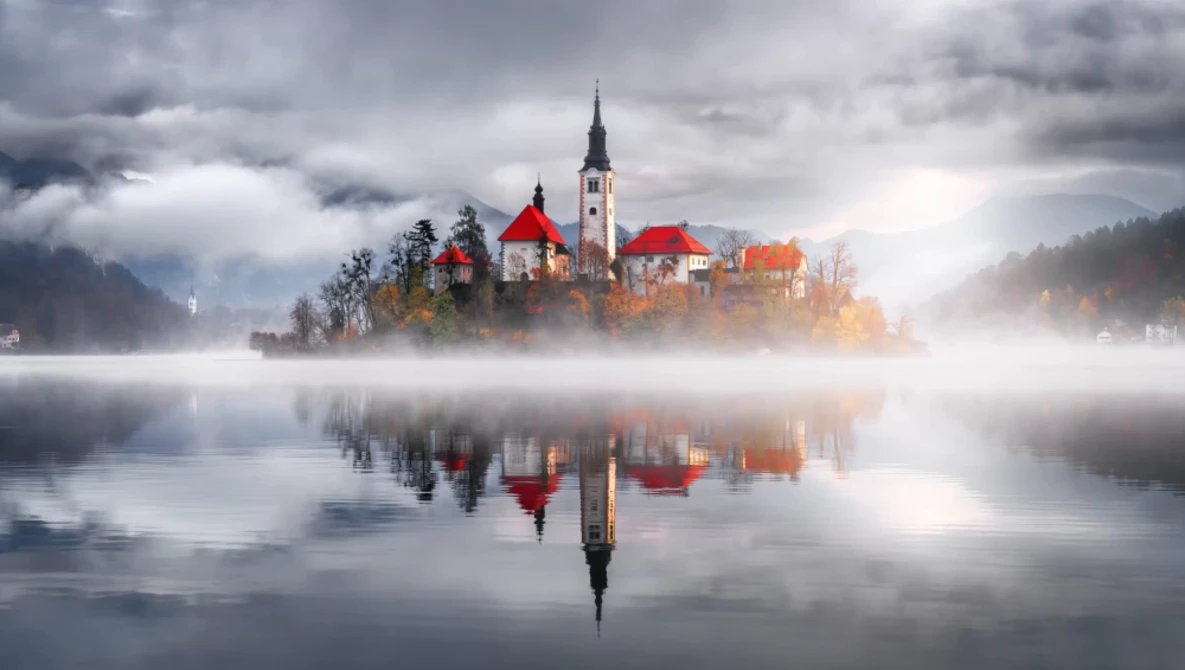 Church with red roof on misty island reflected in still water at dawn.