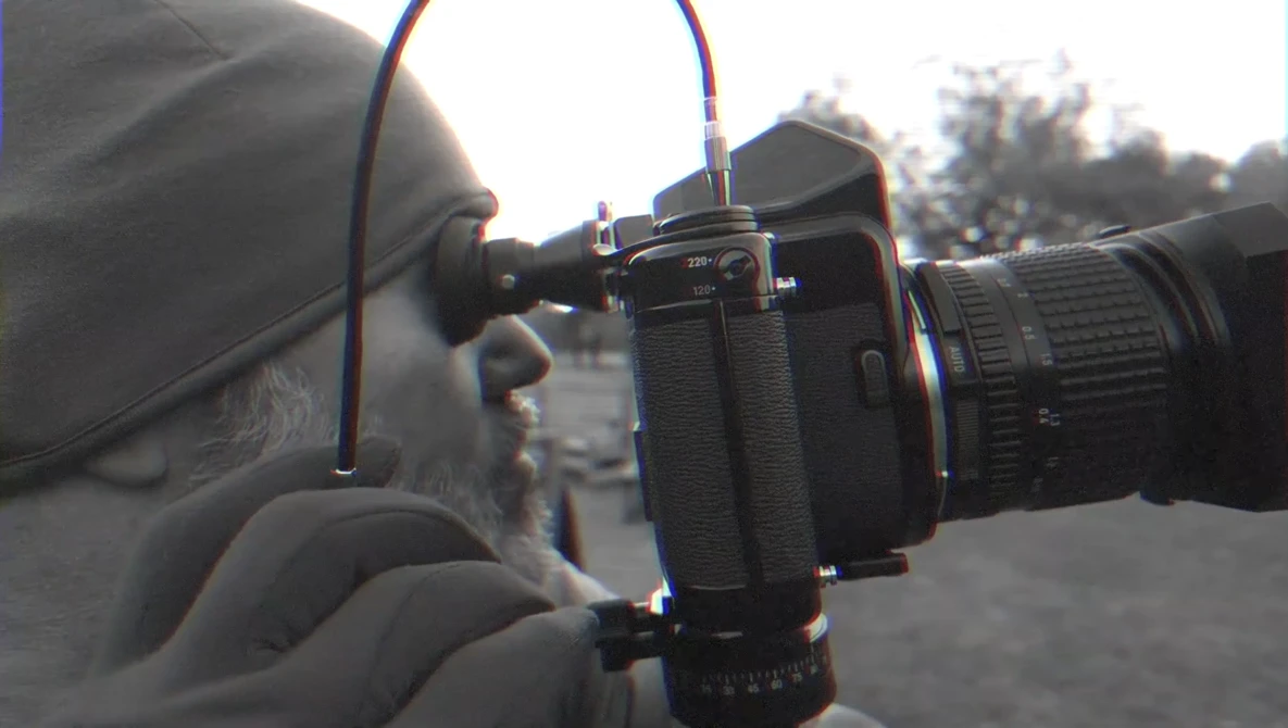 Photographer holding a camera with telephoto lens up to their eye, viewed from the side profile outdoors.