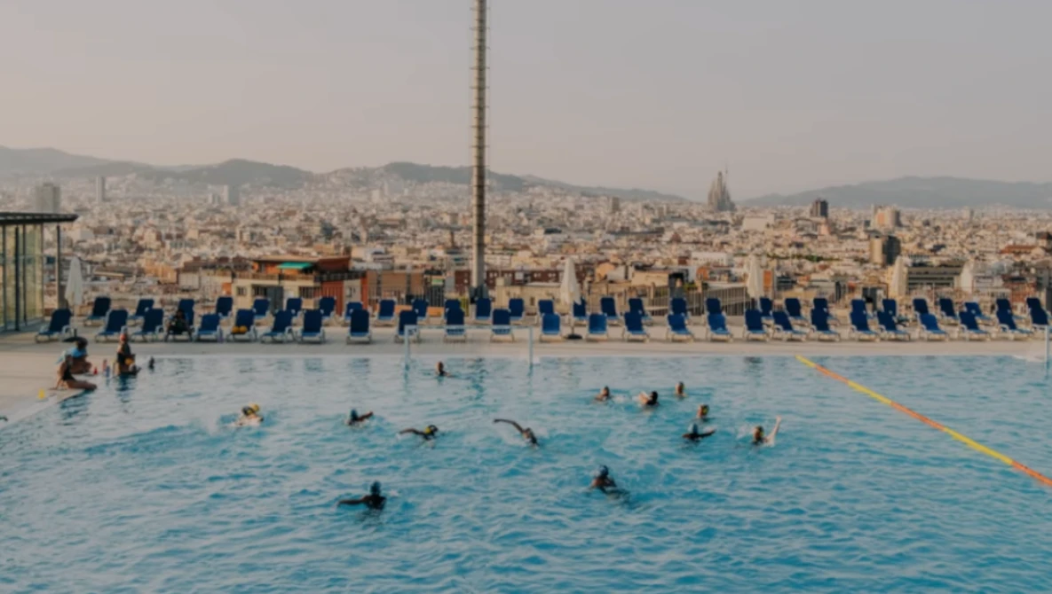 Swimmers in a bright blue public pool with a dense cityscape and mountains visible in the background.