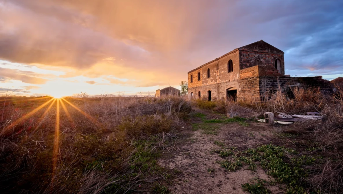Abandoned stone building at sunset with golden light rays streaming across overgrown fields.